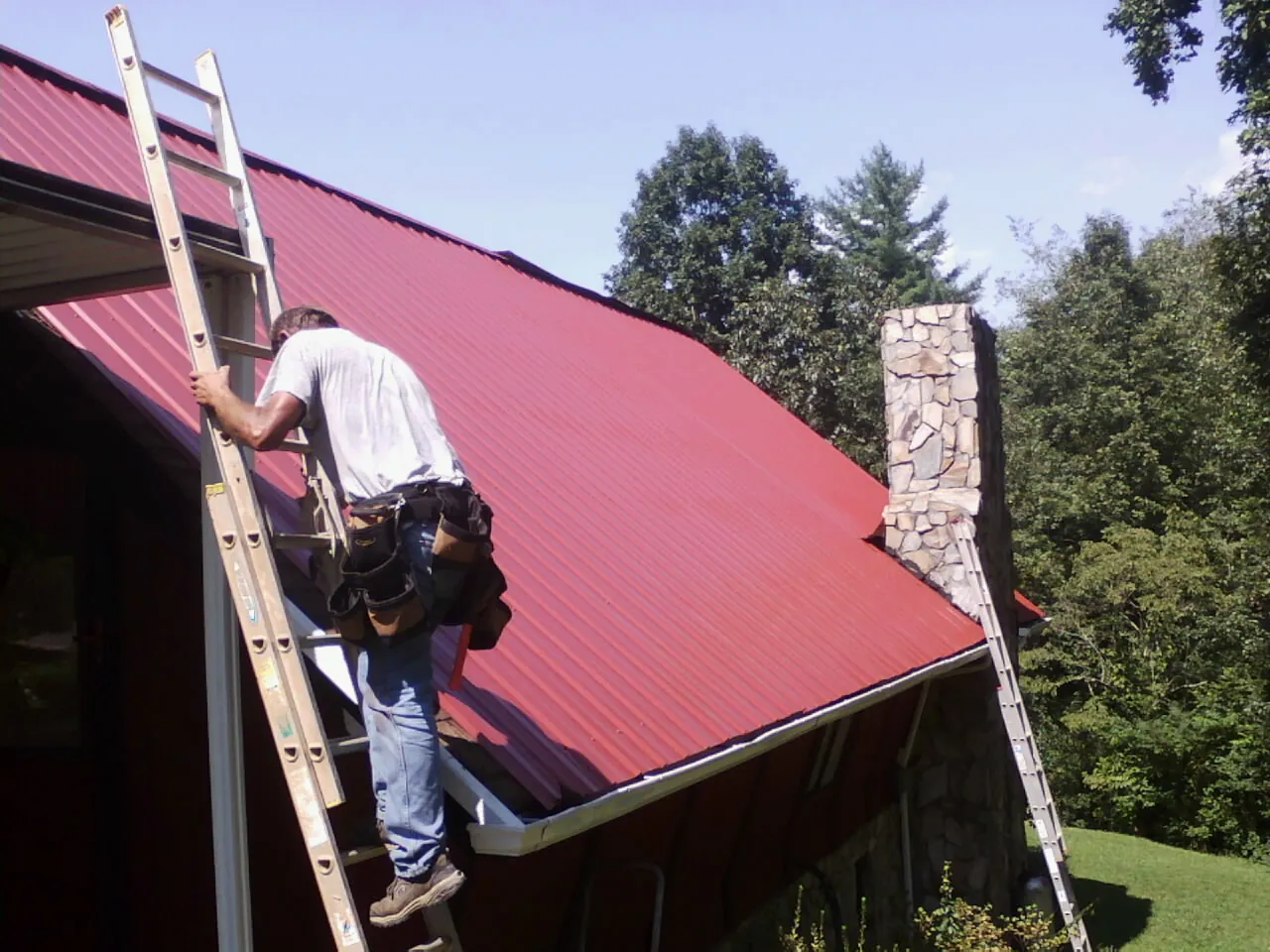 Roofer on a ladder beside a red metal roof and stone chimney — Soffit & Fascia Repair by Roofing Brothers in Mount Wolf